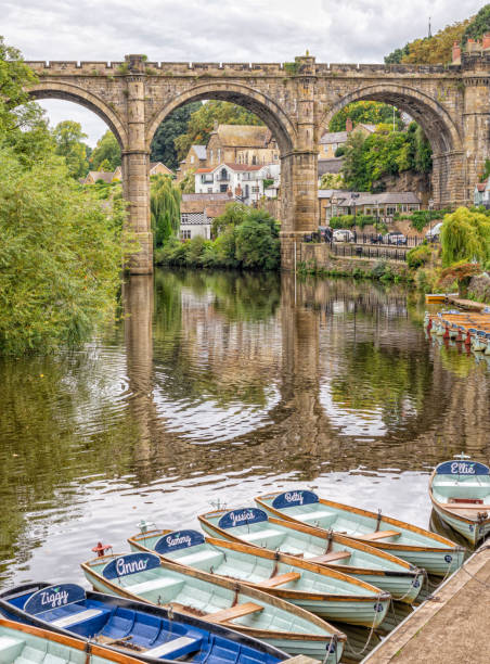 An historic arched viaduct stretches across a river and rowing boats are moored in the foreground. The arches are reflected in the water.