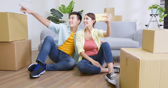 asian couple sitting on the floor use smartphone having video call with friends and family happily in new house with many boxes