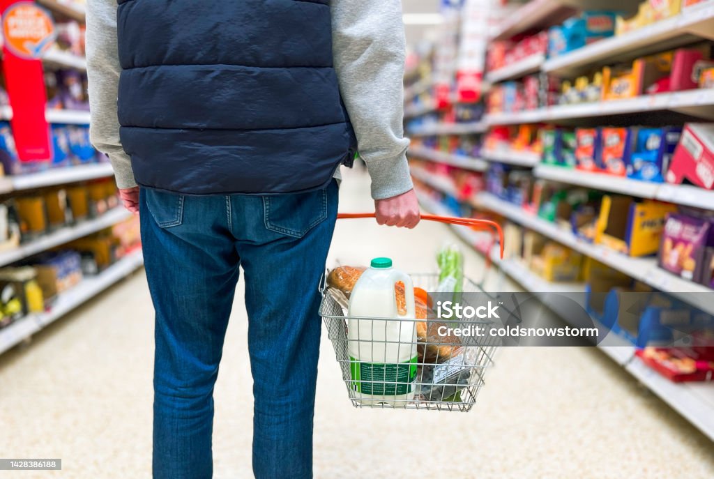 Man holding shopping basket with bread and milk groceries in supermarket Low angle close up color image depicting a man holding a shopping basked filled with essential fresh groceries like bread and milk in the supermarket. Inflation - Economics Stock Photo