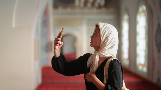 A young female tourist is visiting a mosque and taking photos and videos with her smart phone.