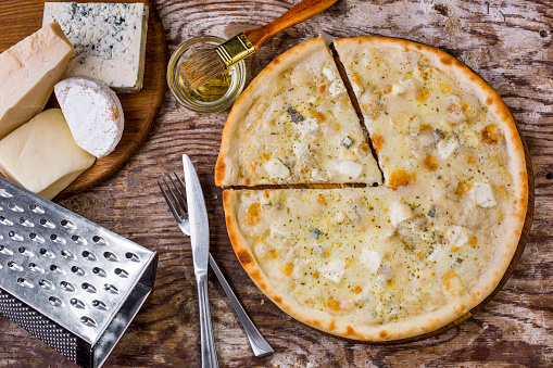 Pizza with different types of cheese on a wooden table with a grater, cutlery and four types of cheese lying sideways on a wooden board