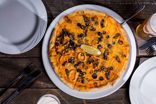 Pizza with mussels, squid rings, olives, lemon and cheese in a plate on a wooden table with cutlery glass of beer