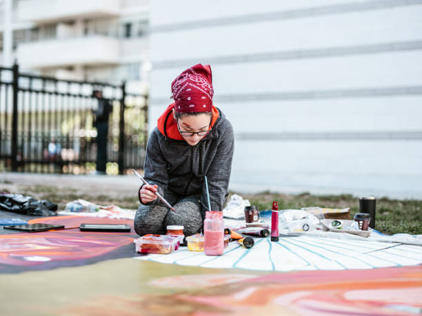 Young Latin woman creating outdoor mural on the lawn. She is dressed in casual outfit, wearing bandana and eyeglasses. Exterior of public park in the city.