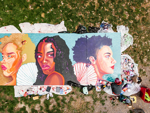 Young Latin woman creating outdoor mural on the lawn. She is dressed in casual outfit, wearing bandana and eyeglasses. Exterior of public park in the city. Captured by drone from above.