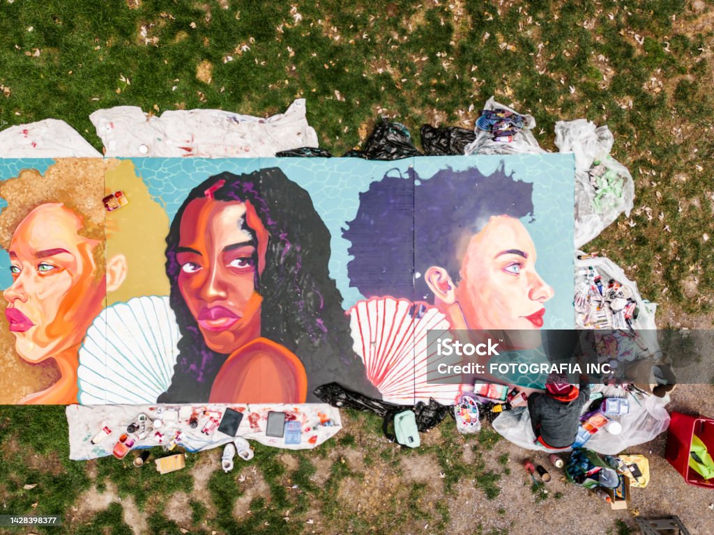 Drone view of Young Latin woman creating outdoor mural Young Latin woman creating outdoor mural on the lawn. She is dressed in casual outfit, wearing bandana and eyeglasses. Exterior of public park in the city. Captured by drone from above. Mural Stock Photo