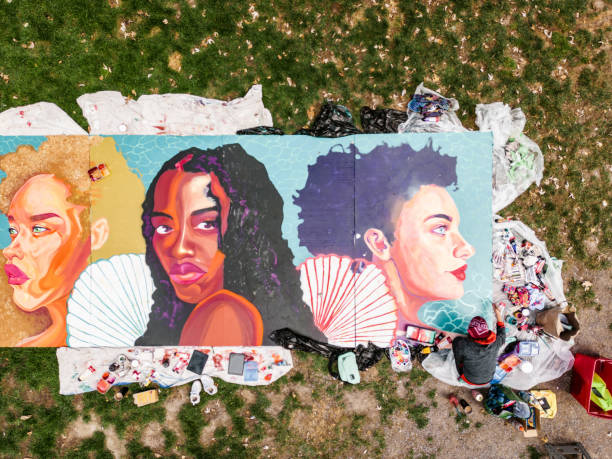 Drone view of Young Latin woman creating outdoor mural Young Latin woman creating outdoor mural on the lawn. She is dressed in casual outfit, wearing bandana and eyeglasses. Exterior of public park in the city. Captured by drone from above. local artist stock pictures, royalty-free photos & images