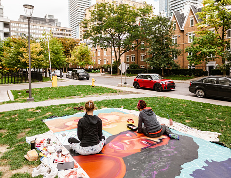 Two Young female artists creating outdoor mural on the lawn. They are  dressed in casual outfit. Exterior of public park in the city.