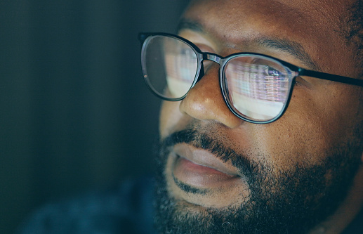 Close-up stock image of an afro-caribbean man watching a computer screen of numbers which are reflected in his glasses.