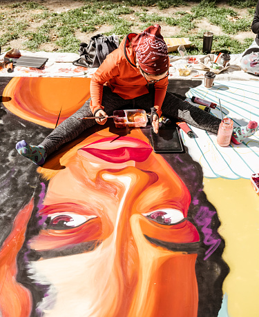 Young Latin woman creating outdoor mural on the lawn. She is dressed in casual outfit, wearing bandana and eyeglasses. Exterior of public park in the city.