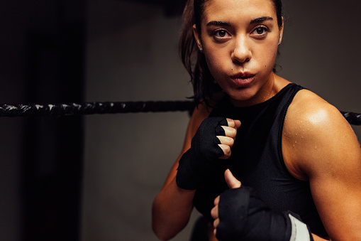Athletic woman standing in a boxing ring with wrapped hands before a fight. Female boxer having a training session in a boxing gym.