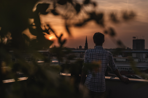 Silhouette of man watching sunset on rooftop garden. Male standing on apartment balcony terrace and looking at Novi Sad skyline at sunset. Person enjoys evening on green roof during dawn in Serbia.