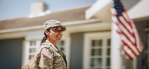 Happy female soldier looking away with a smile while standing outside her house with her bag. American servicewoman coming back home after serving her country in the military.