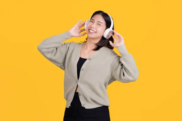 Photo of Happy asian beautiful woman listening to music She is wearing wireless headphones and standing isolated on a bright yellow background.