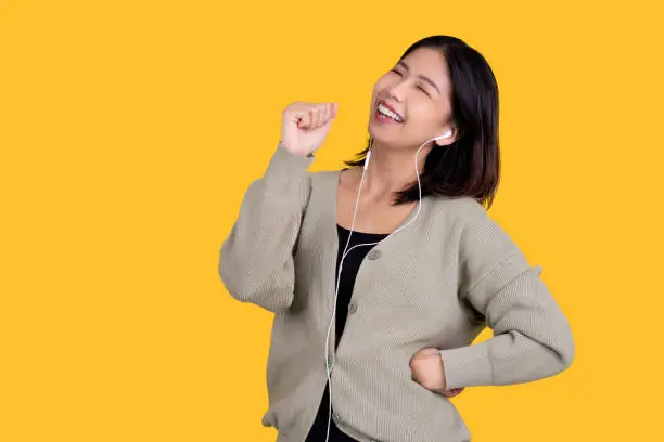 Photo of Happy Asian beautiful woman singing her favorite song. She is wearing white headphones isolated on a bright yellow background.