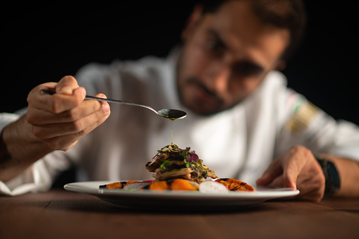 A male chef pouring sauce from the spoon on meal on a black background.