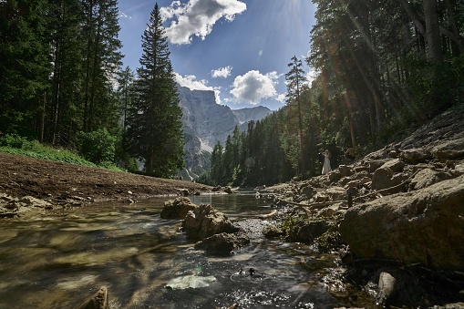 a rocky mountain and stream flows in a valley, a mountain stream running through a forest under a partly cloudy sky