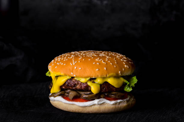 hamburger con panino al sesamo, insalata, salsa gialla, cotoletta, cetrioli sottaceto, funghi, maionese su una tavola rotonda di legno nero su sfondo scuro. orientamento orizzontale - symmetry burger hamburger cheese foto e immagini stock