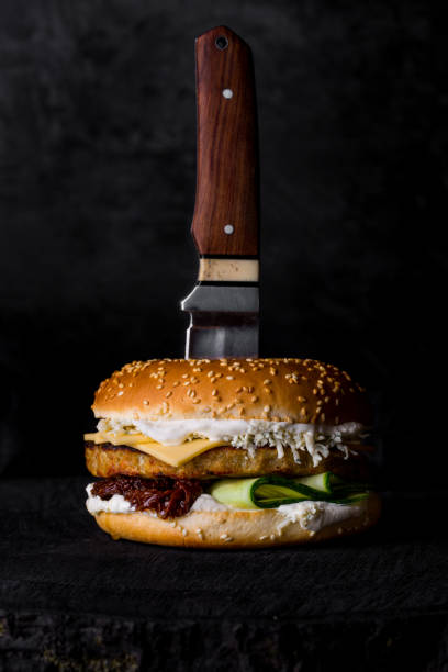 istock Burger with sesame bun, cutlet, cheese, cucumbers, grated cheese, mayonnaise, pickled vegetables on a black wooden board on a dark background a knife inserted in the center of the bun. Vertical orientation 1428421821