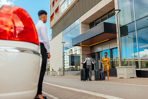 A stylish African American couple is leaving the luxurious hotel they just spent their weekend at. They are carrying their luggage with them as the limo driver is waiting for them next to the car.