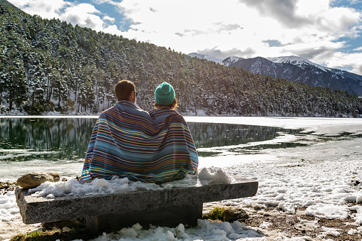 Back view of a man and woman couple covered with a tribal blue blanket sitting on a stone bench while are looking at a beautiful icy lake surrounded of snowy forest and mountains. Winter landscape.