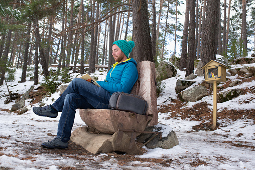 Attractive man sitting on a wood bench while is reading a book on a snowy forest. Bookcroosing concept.