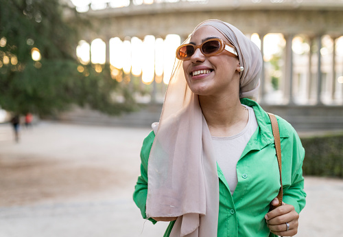 Young arab girl smiling outdoors, wearing sunglasses, strolling at sunset while traveling.