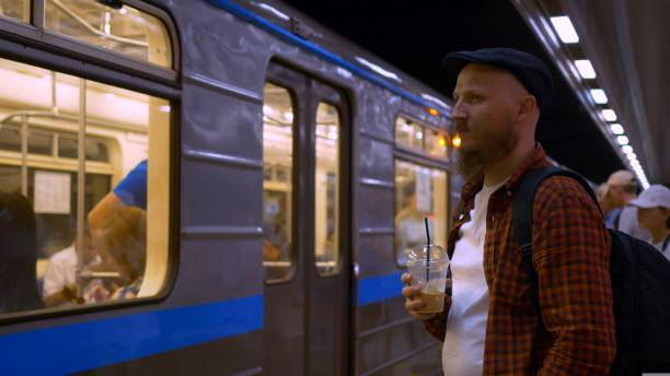 istock A man with a beard in a cap is waiting for a subway train, drinking cold coffee with ice. A wagon pulls up to the underground subway platform. The headlights of a subway train illuminate a man. 1428432452