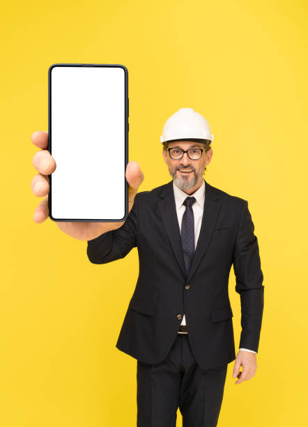 istock Holding smartphone middle aged architect businessman wearing black suit and construction helmet isolated on yellow background. Businessman in suit construction hard hat and eye glasses 1428437761
