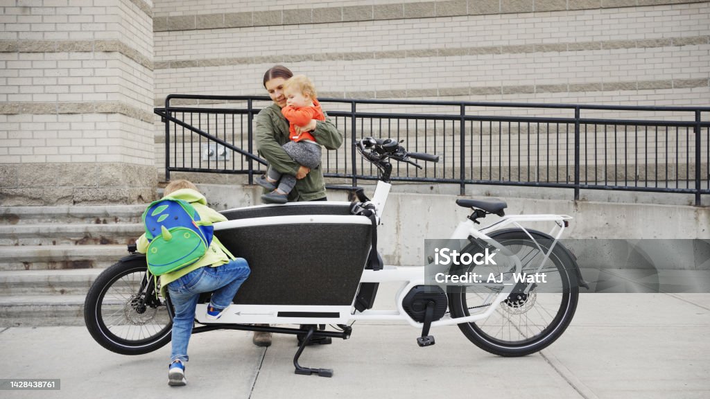 Mother and little sons getting off a cargo bike on a sidewalk Mother and her two cute little sons getting off a parked cargo bicycle at the entrance to a school Cargo Bike Stock Photo