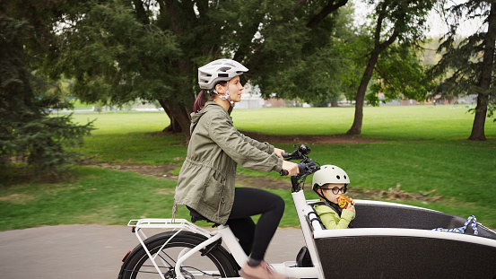 Mother riding a cargo bike along a path in a park with her young son sitting in the front cart