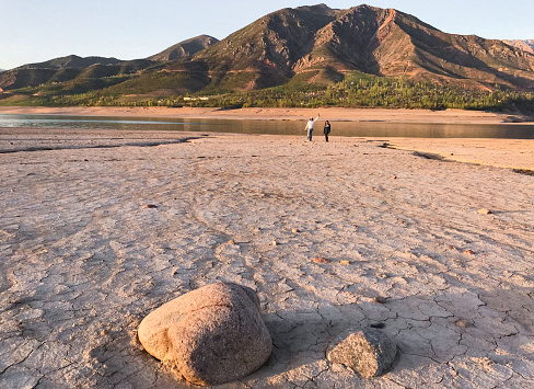 Beautiful landscape with setting sun: mountains, lake, cracked earth with stones -dried-up riverbend. Two people are standing in the distance.