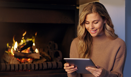 Fireplace and happy woman on digital tablet reading and relaxing at home with smile by warm fire. Portrait of a female with a smile for ebooks or entertainment with touchscreen technology and wifi