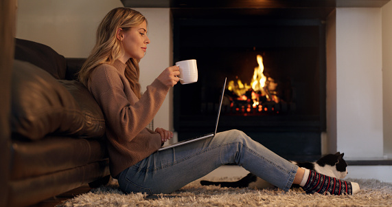 Happy woman, fireplace and laptop while browsing the internet, watching movie or writing article with coffee for a blog at home. Young female streaming online film or series subscription drinking tea