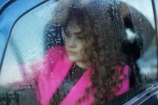 Sad woman looks out foggy car window. Window with rain drops. Curly-haired woman is riding in car in pink jacket