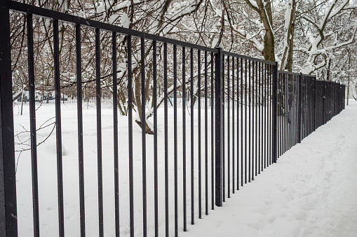 A long black transparent metal fence encloses the park in winter during snowfall. Bad weather. Diagonal arrangement.