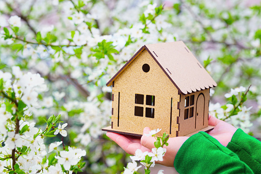 Female hands holding a wooden house model under a blooming tree. Spring home selling season concept.