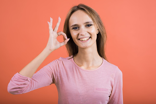 A young Caucasian girl in a pink jumper smiles and shows an Okay gesture isolated on a red studio background.