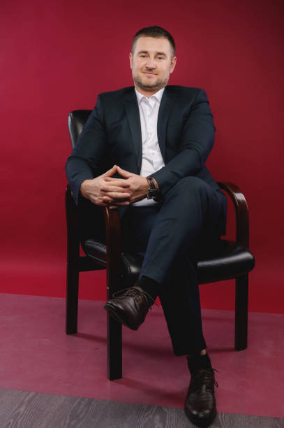 ritratto di un uomo d'affari in un abito nero seduto in uno studio su una sedia contro un muro vuoto - presidente di organizzazione foto e immagini stock