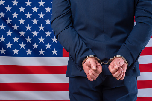 Business man in suit jacket handcuffed with united states flag in background. Political and financial corruption in America