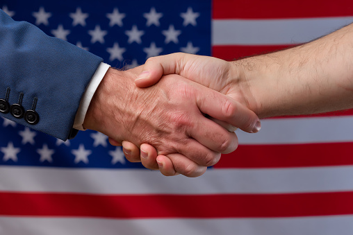Two men shaking hands with the United States flag in the background. Cooperation and political protection of citizens in America. Alliance with the USA