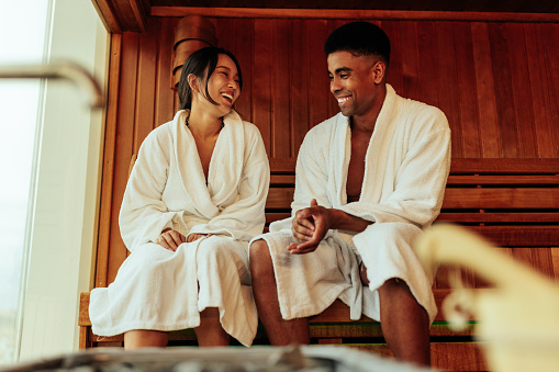 A young ethnically diverse couple is in the sauna. They are sitting in bathrobes, talking to each other and smiling.