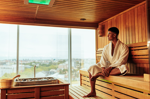 A young African American man is wearing a bathrobe and enjoying the view of the city in the sauna.