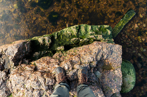 Feet of a man in rough trekking shoes standing on a coastal stone. Travel lifestyle vertical photo