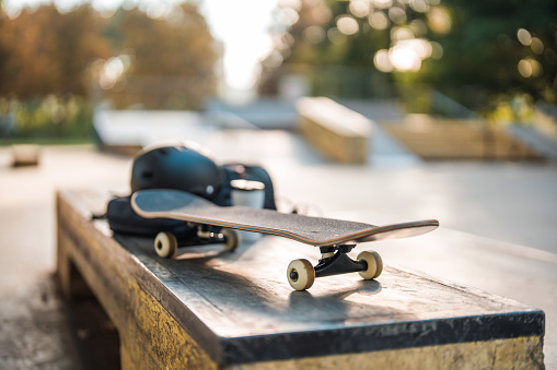 Skateboarding items on top of the curb box at a skatepark. Portable plastic coffee cup next to a protective helmet. Focus on the board. Blurred background with other ramps visible.