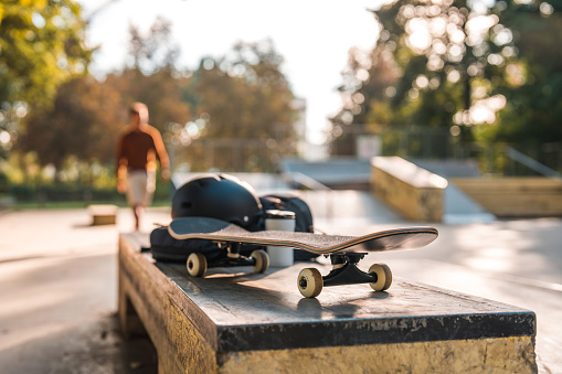 A new skateboard on top of the curb box at a skatepark. Portable plastic coffee cup next to a protective helmet. Focus on the board. Blurred background with other ramps visible and a young person approaching.
