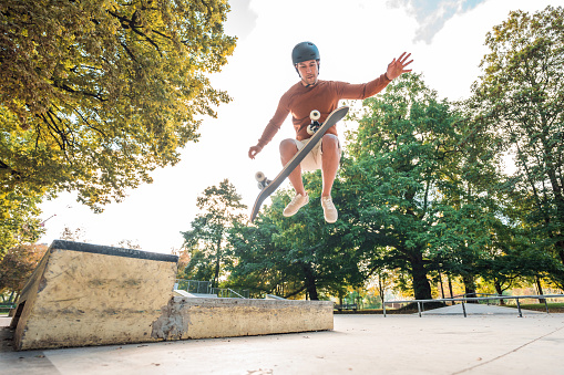 Male skateboarder of Asian descent, arms spread wide  apart while trying a kick-flip  with his skateboard. Full length image. Trees in the background.