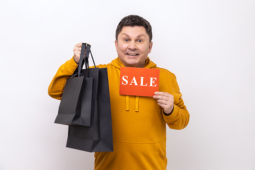 Portrait of satisfied man holding paper bags and sale sign, shopping and discounts, looking at camera with happy face, wearing urban style hoodie. Indoor studio shot isolated on white background.