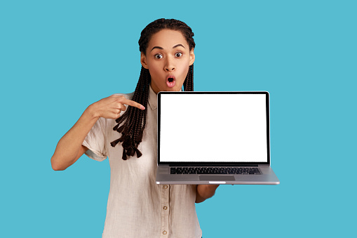 Impressed scared woman with dreadlocks opens mouth from interest and thrill, points at laptop with empty screen, feels fascinated, wearing white shirt. Indoor studio shot isolated on blue background.