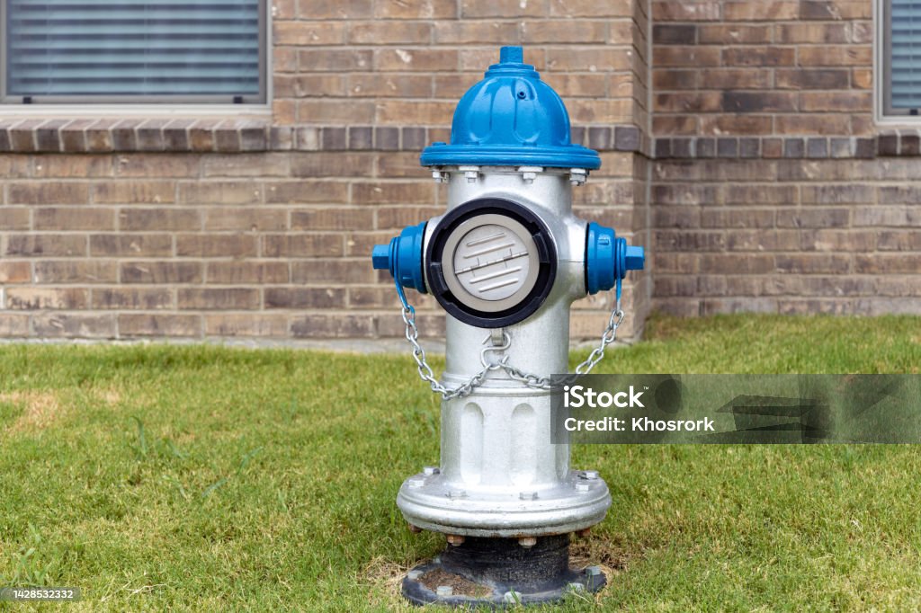 Grey and blue fire hydrant on the grass int he yard in residential complex with brick wall of the building on background. Accidents and Disasters Stock Photo