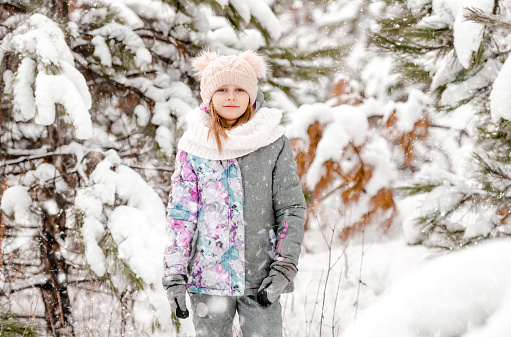 Pretty child girl in winter forest with snow trees looking at camera. Preteen female kid wearing hat in cold weather portrait